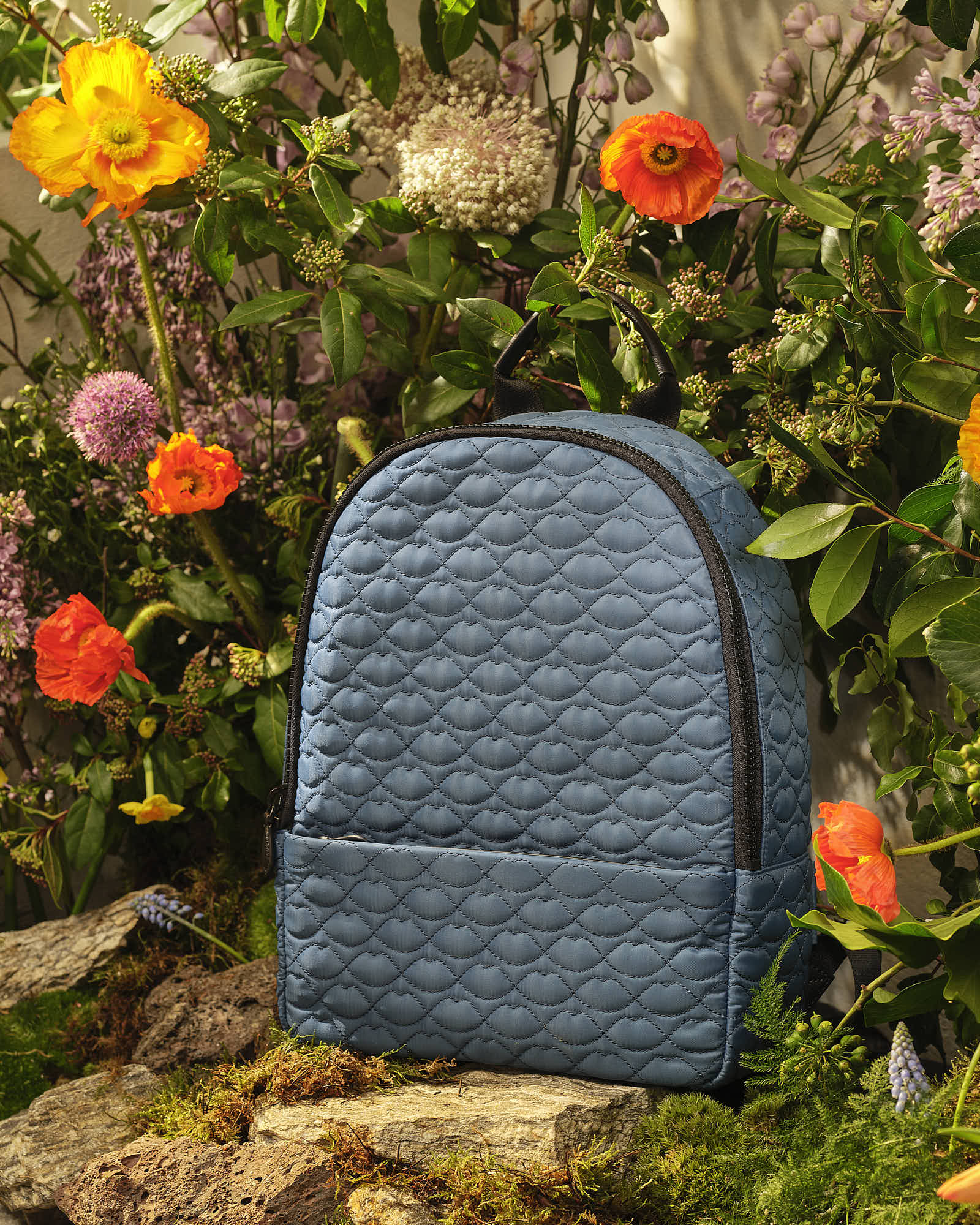 Blue quilted backpack surrounded by vibrant flowers and greenery.