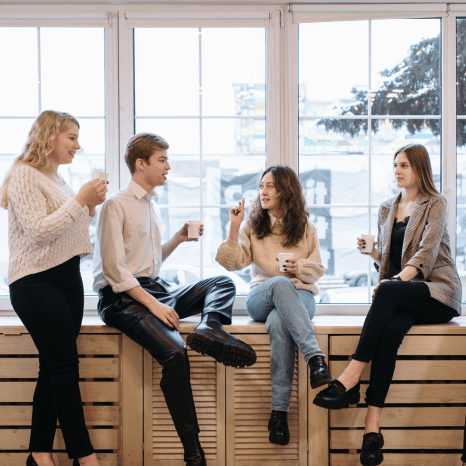 A group of four young people, likely friends or colleagues, engaged in conversation and enjoying drinks together in what appears to be a casual office or workplace setting. Their relaxed and friendly demeanor suggests a positive and collaborative work environment.