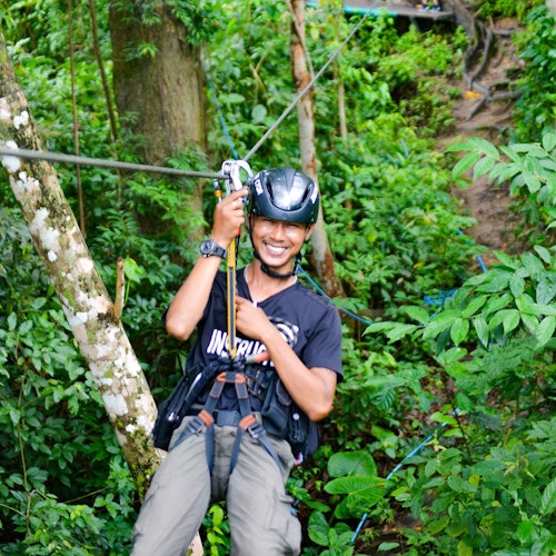 一位人士佩戴头盔和安全带，在郁郁葱葱的绿色森林中进行高空滑索时微笑。