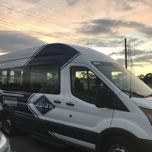 En vit passagerarbuss märkt "Gray Line" med "Sightseeing Everywhere" på sidan, parkerad under en molnig solnedgångshimmel.