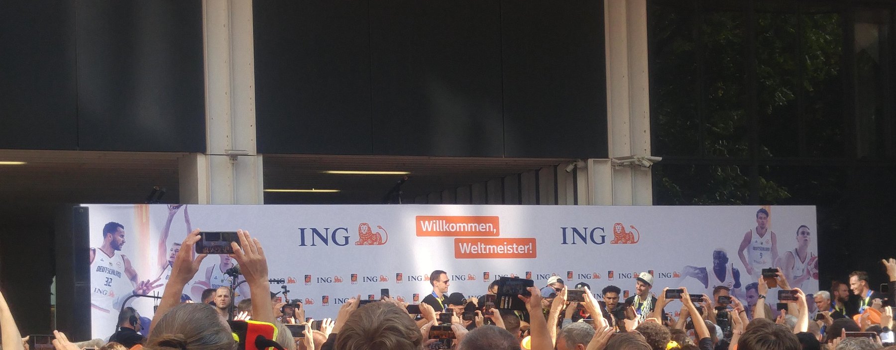 Germany’s basketball world champions at a reception beneath a welcome banner, with spectators raising phones in the foreground.
