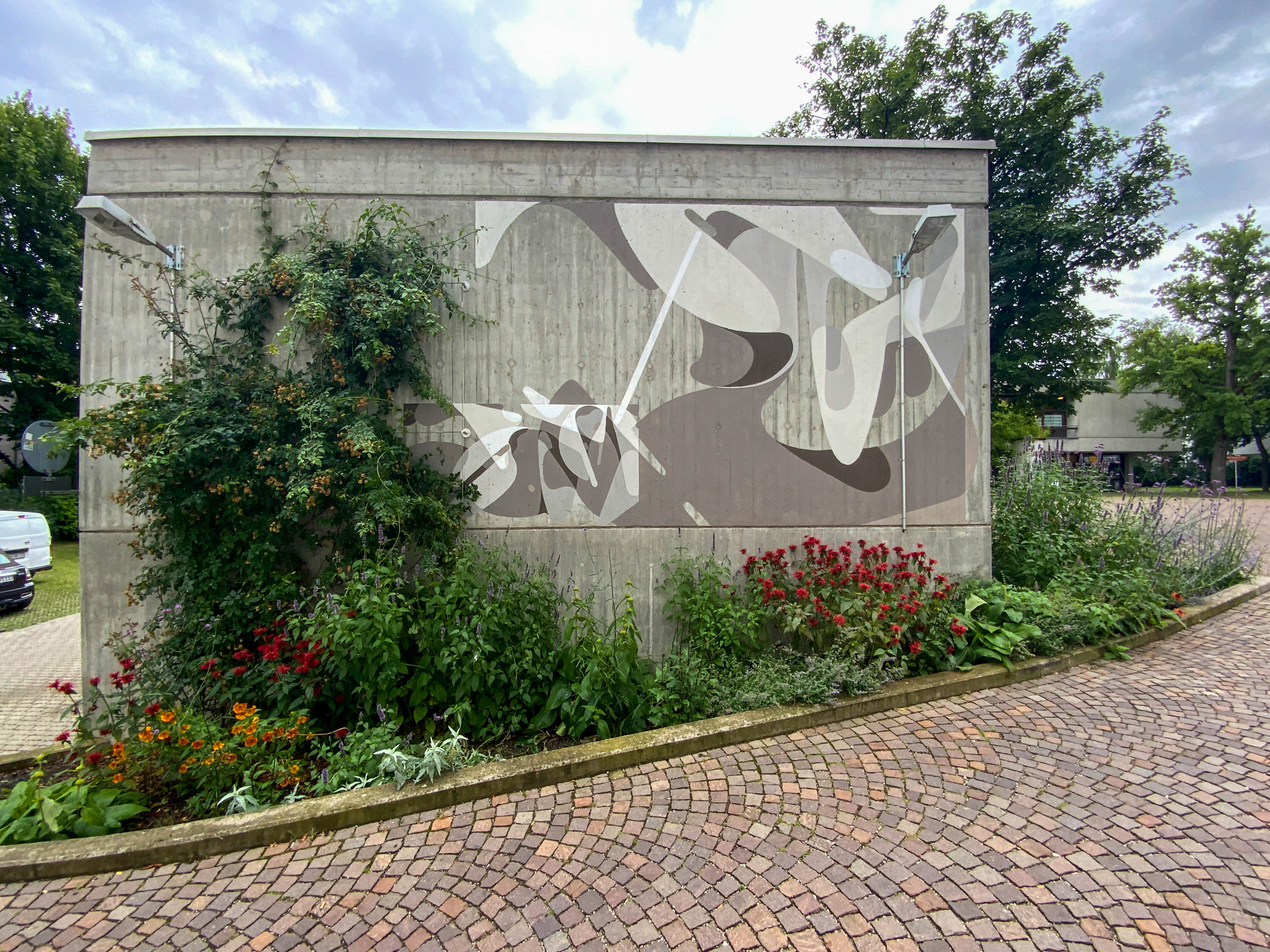 An abstract monochrome mural painted on a gray wall sourrunded by green plants and red flowers beside a pathway.