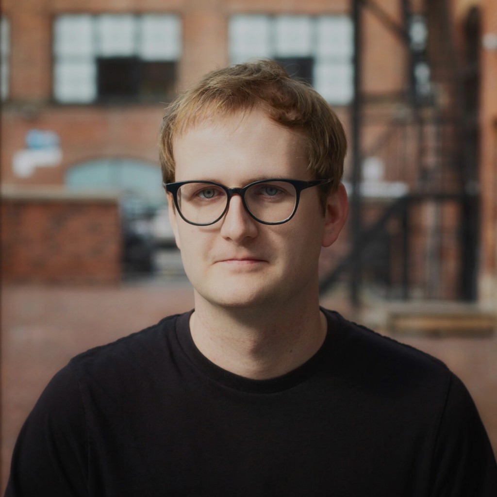 A man with glasses and a black t shirt standing in front of a brick wall