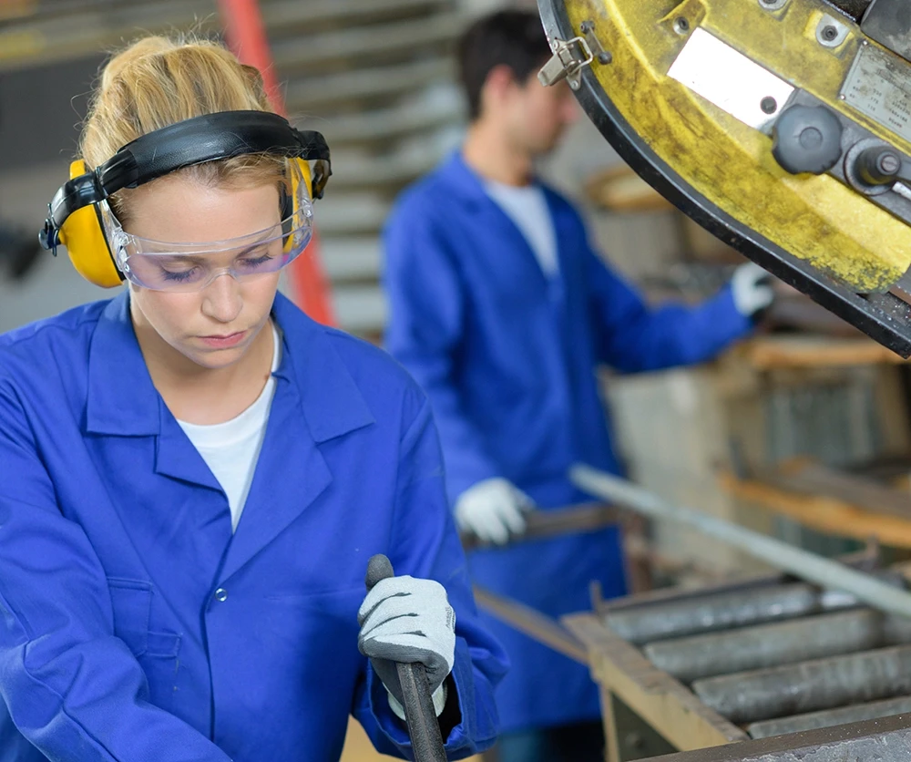 Two workers in blue overalls are operating machinery in a workshop, focusing on their tasks. Safety equipment is visible.