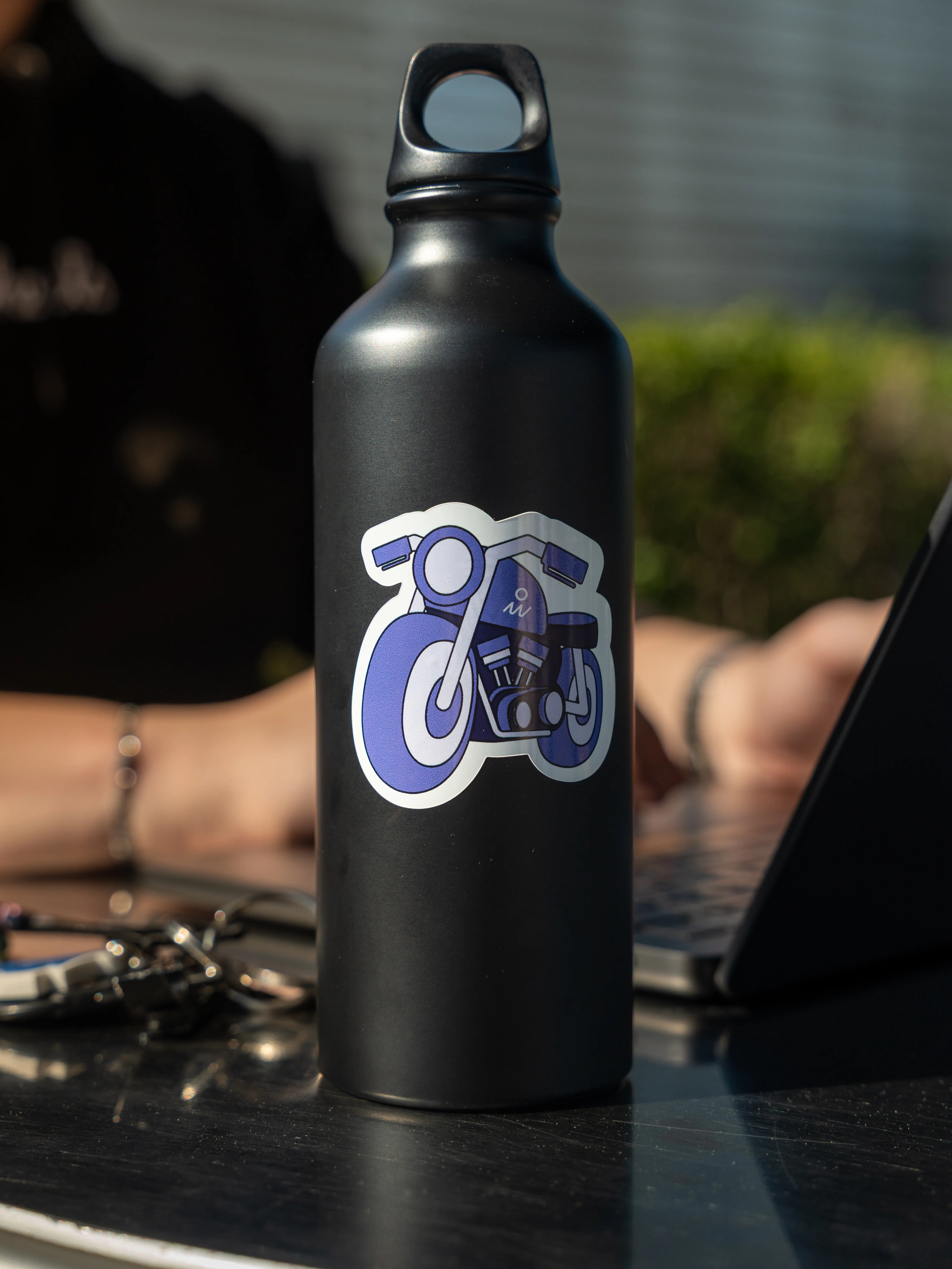 Gourde noire avec sticker moto On Wheels posée sur une table, univers motard