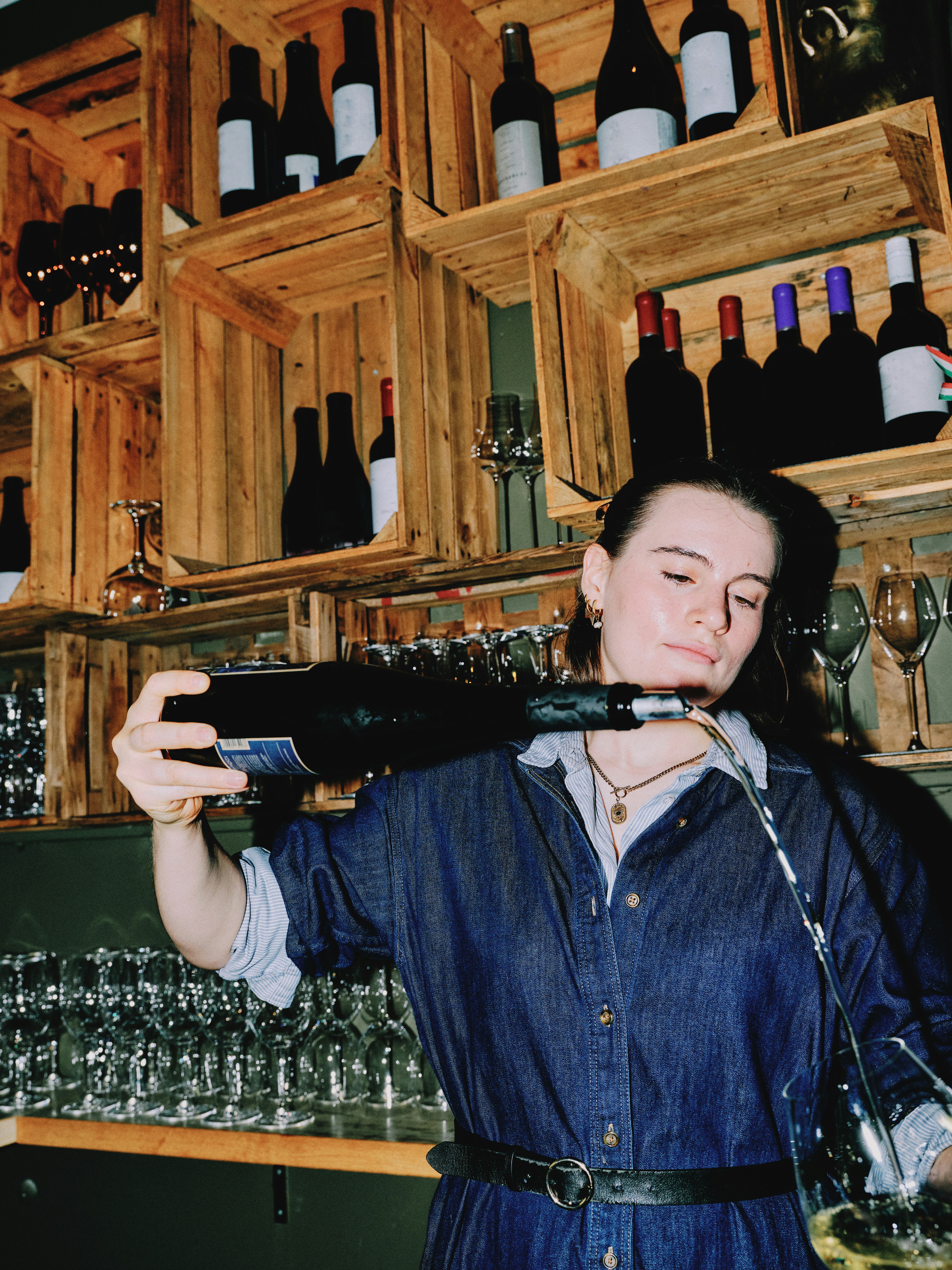 Female bartender in a denim dress pouring wine dramatically overhead at a rustic wine bar, wooden crate shelves and glassware in the background