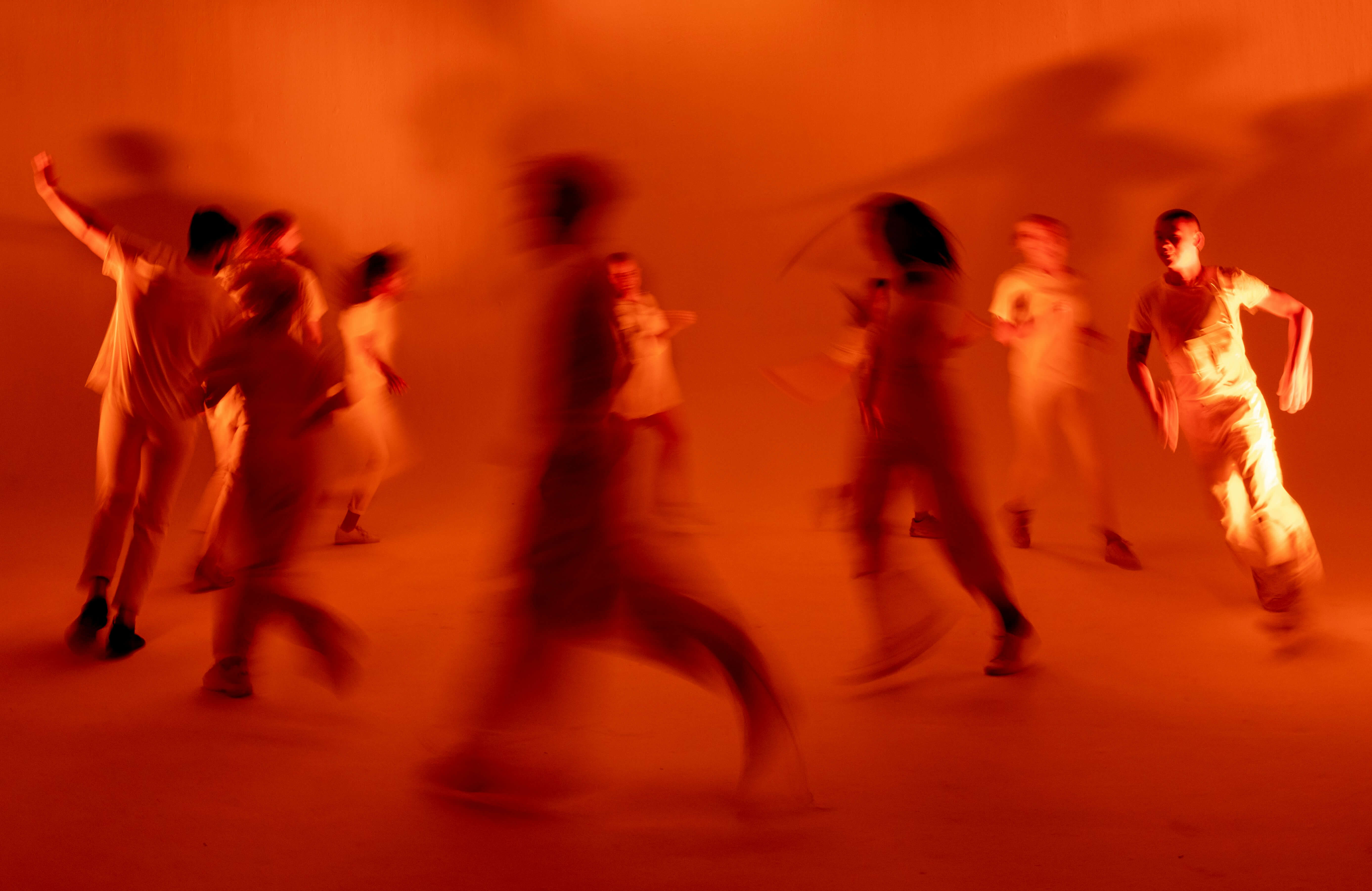 Artistic photo of people in motion, captured with a slow shutter speed that blurs their figures against a glowing orange background. The scene conveys energy and movement, with dramatic lighting casting elongated shadows and adding intensity to the atmosphere.
