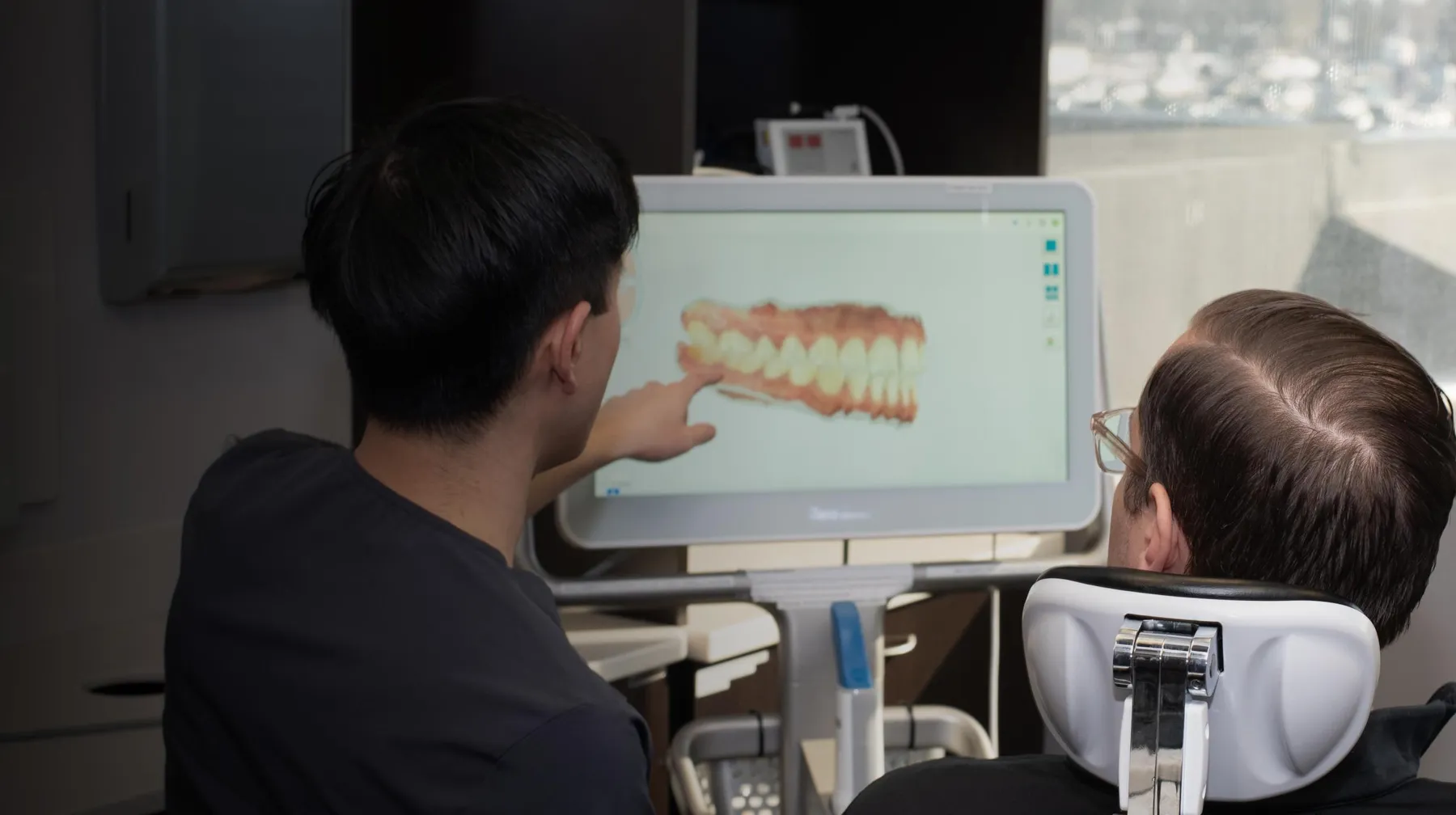 Dentist showing a digital 3D model of teeth on a monitor to a patient seated in a dental chair.