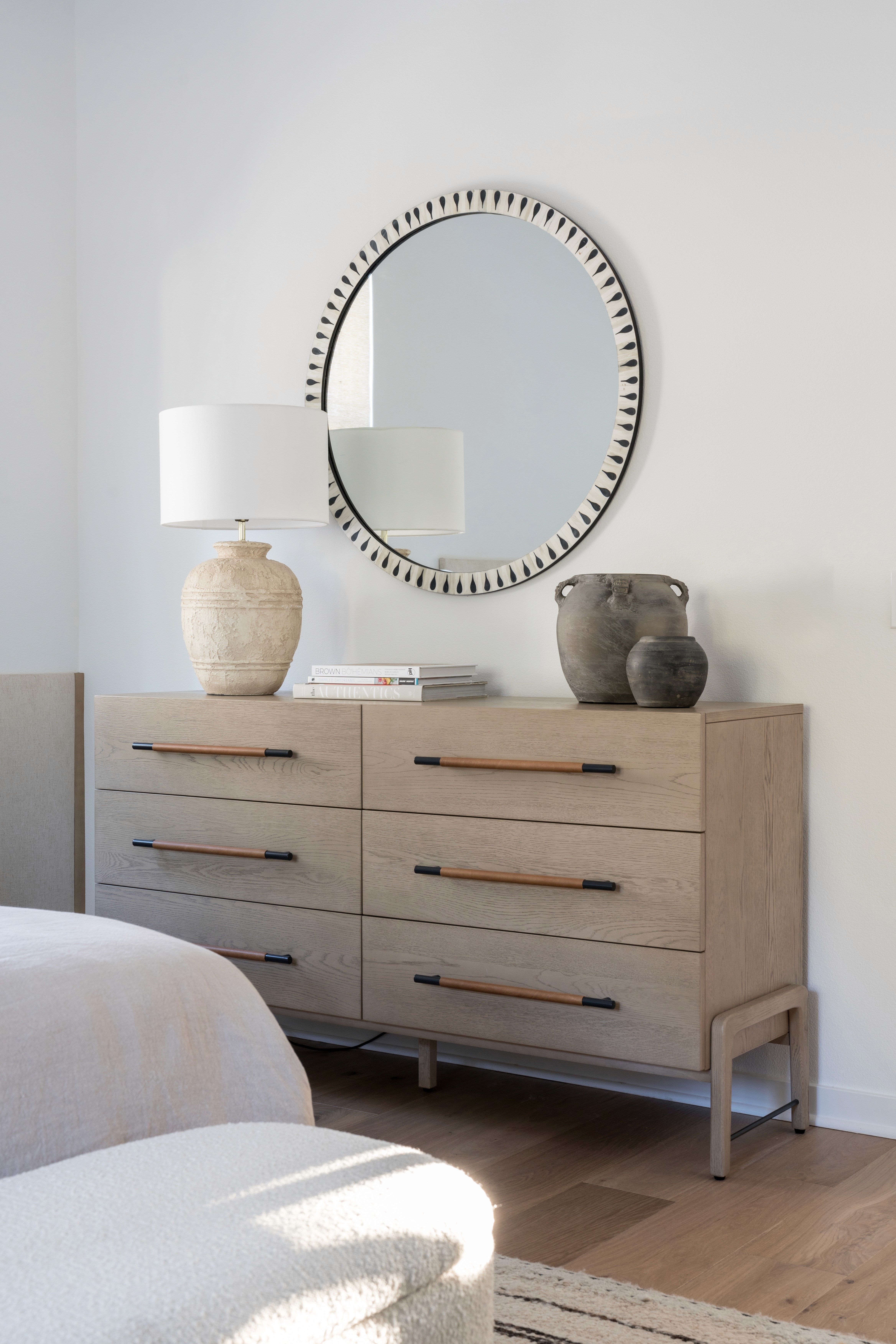 Light oak 6-drawer dresser with round mirror, cream table lamp, and grey antique vases by Jubilee Interiors in Arcadia, California