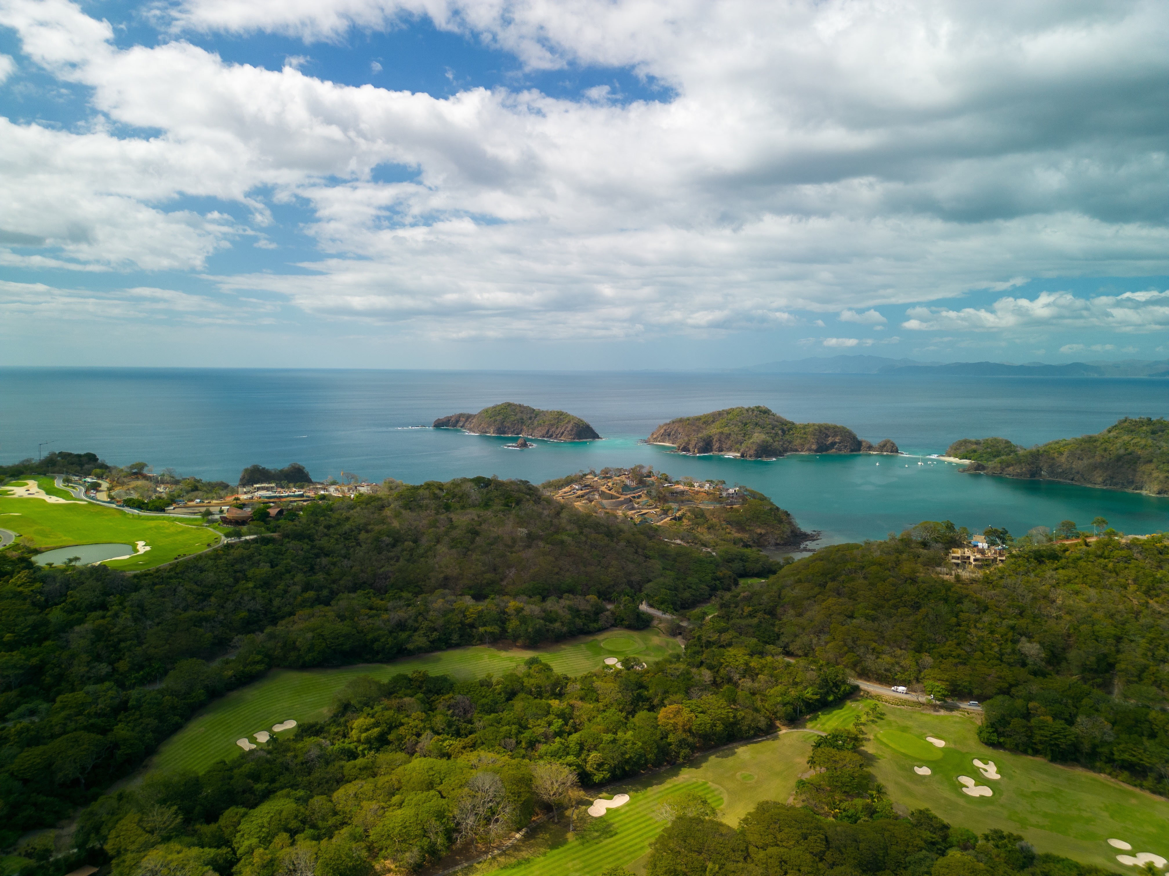 Aerial view of a lush green coastline with a golf course in the foreground, forested hills, and small islands in the ocean under a partly cloudy sky.