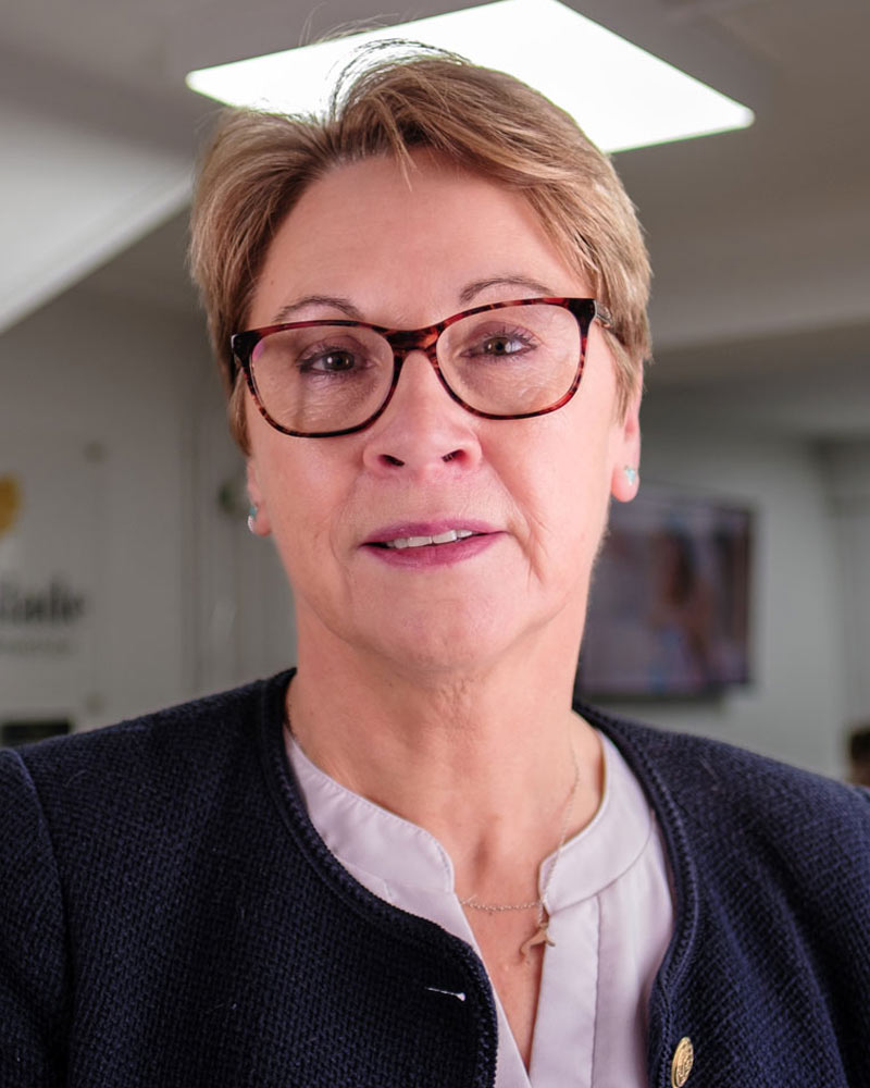 A portrait of Sally, Practice Manager at Cricklade Dental Practice, wearing glasses and a dark blue textured jacket over a light blouse. She is standing in the practice reception area with a blurred logo visible in the background.