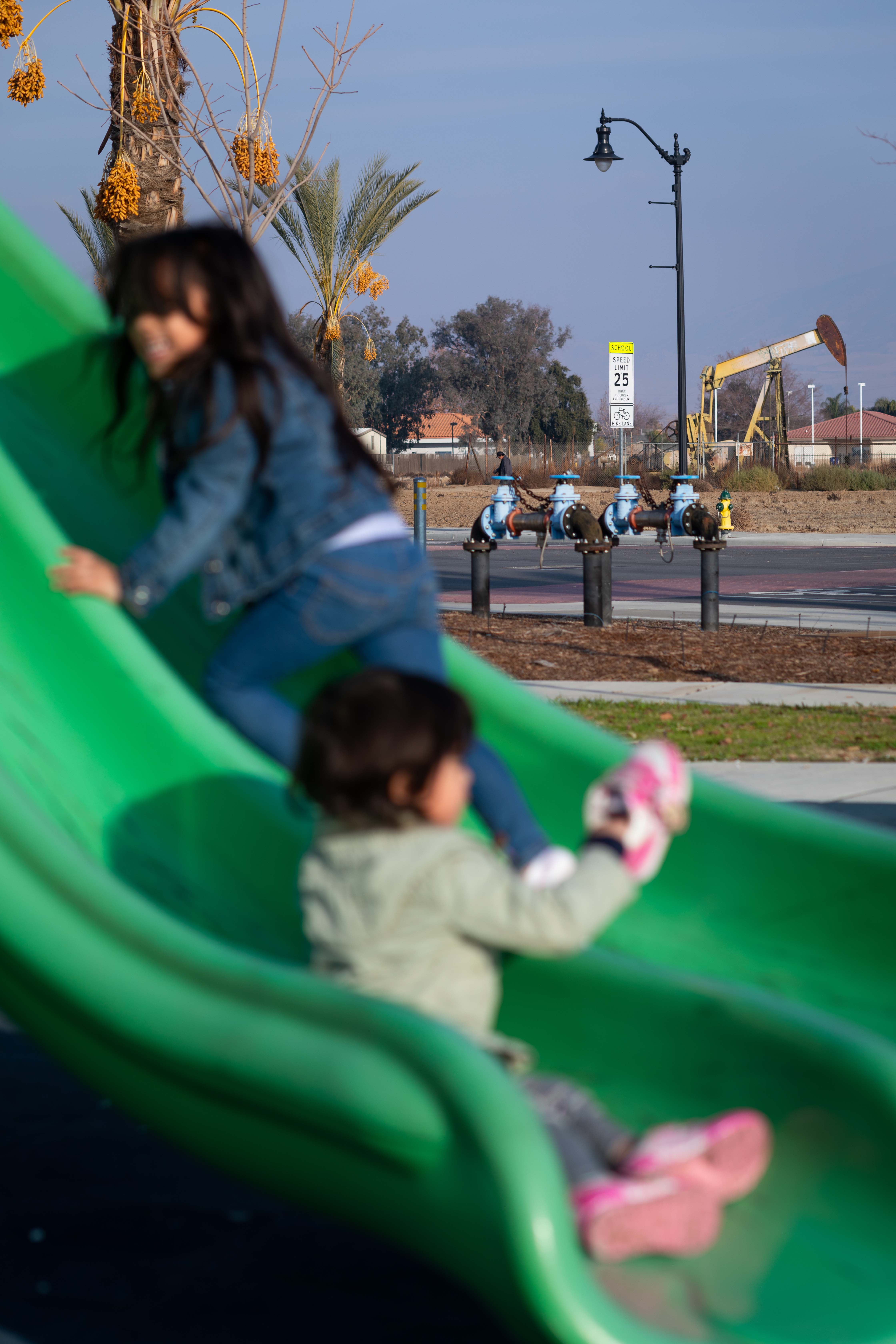 A girl playing on a slide at a park with an oil rig visible in the background.