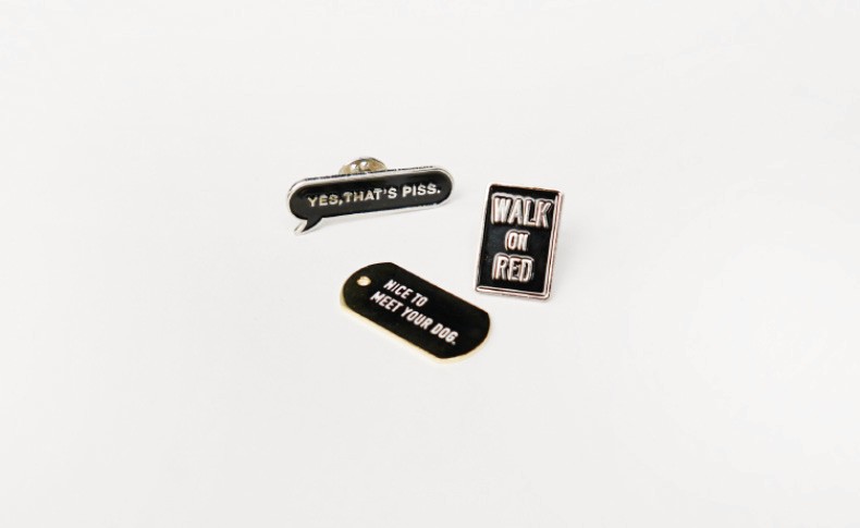 Three black enamel pins are arranged on a white background. The top pin has a speech bubble shape with the text "YES, THAT'S PISS." The middle pin is rectangular and displays "WALK ON RED" in white letters. The bottom pin is oval-shaped and contains the phrase "NICE TO MEET YOUR DOG" in white text.
