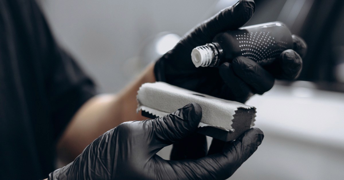 Gloved hands hold a small bottle and pour a substance onto a wrapped applicator pad. A silver car is in the background.