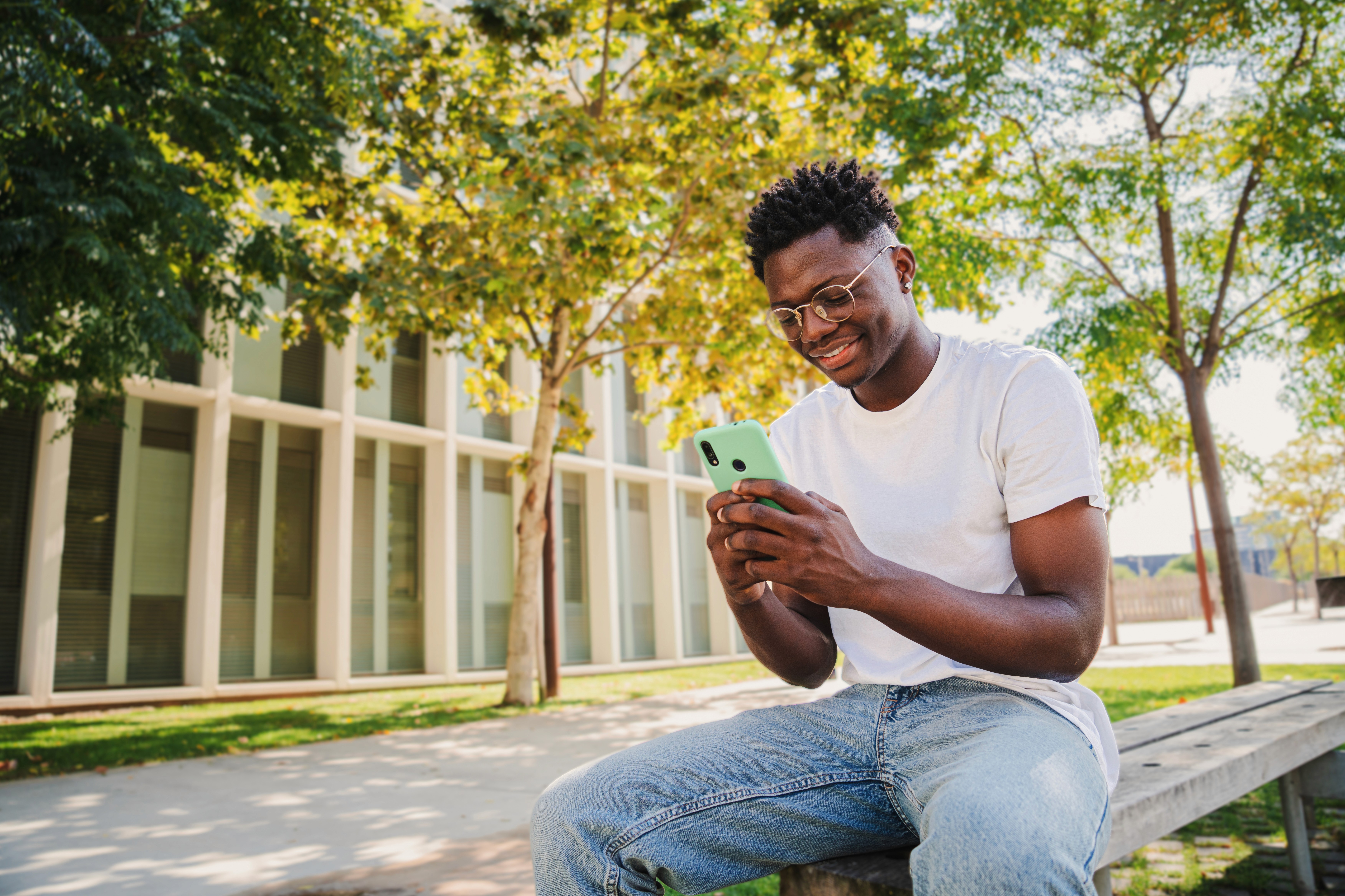 A man wearing a white T shirt and blue jeans uses his cell phone outdoors on a university campus.