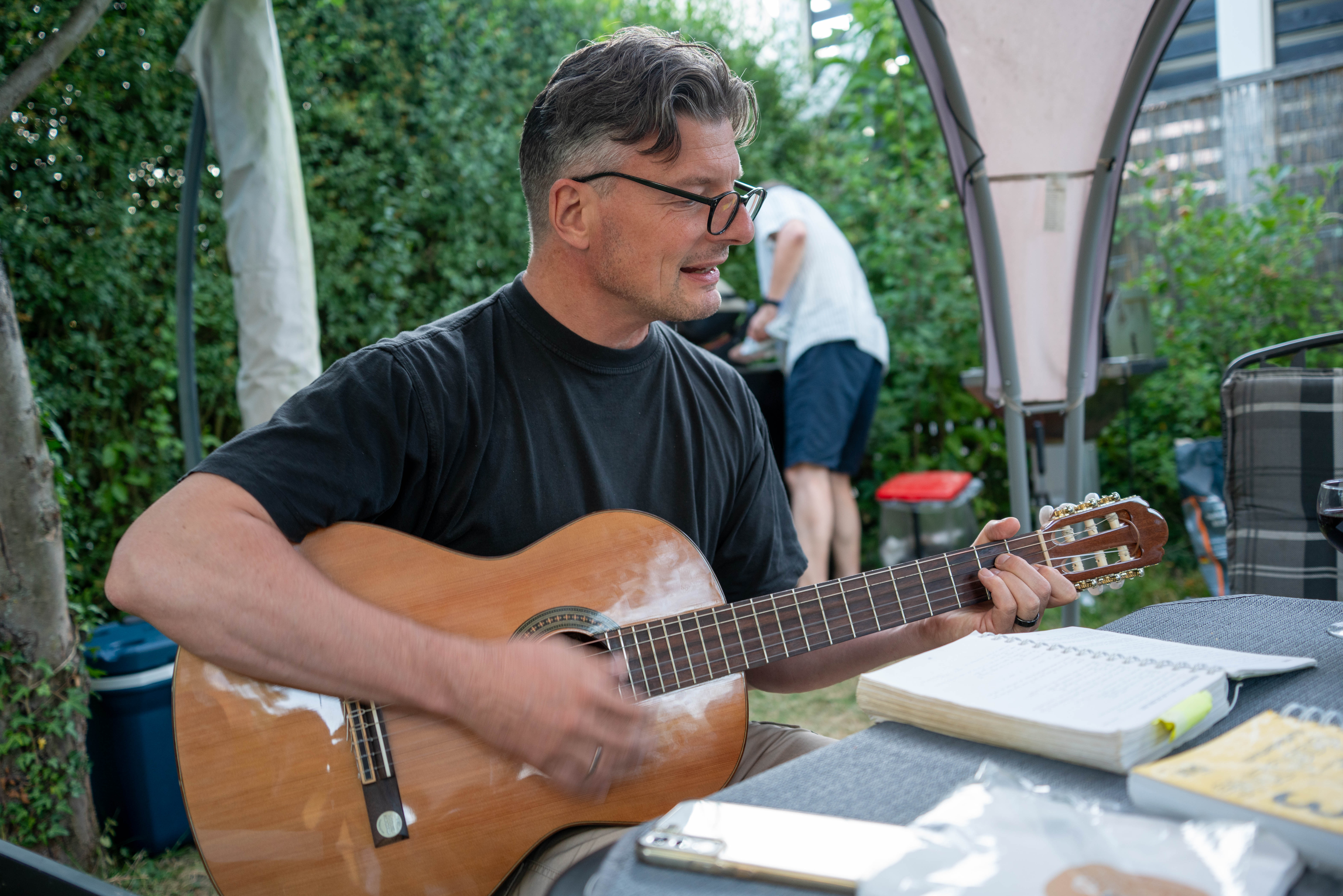 Mann sitzt im Freien und spielt akustische Gitarre, auf dem Tisch liegen Gesangsbuch und weitere Papiere