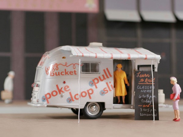 A white model camper van with red and blue lettering reading “poke loopa pull” is parked on a light brown surface. A miniature figure in a yellow dress stands in the open doorway of the van. A chalkboard sign next to the van reads “Learn to knit!” with times listed: 6 to 8 p.m., today, and 3 to 5 p.m., tomorrow. Two small figures, one in white and one in pink, are visible in the background. The van has a striped awning and silver hubcaps.