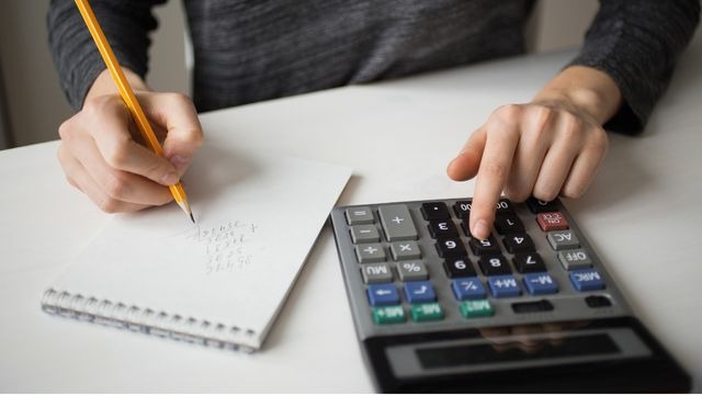 Mãos masculinas anotando números a lápis em um bloco de notas enquanto digita em uma calculadora.
