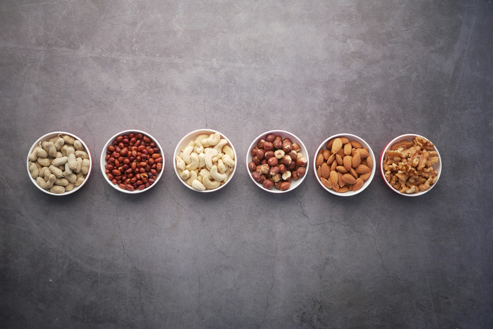 Six small white bowls holding an assortment of nuts and seeds, calorie-dense sources of healthy fats.