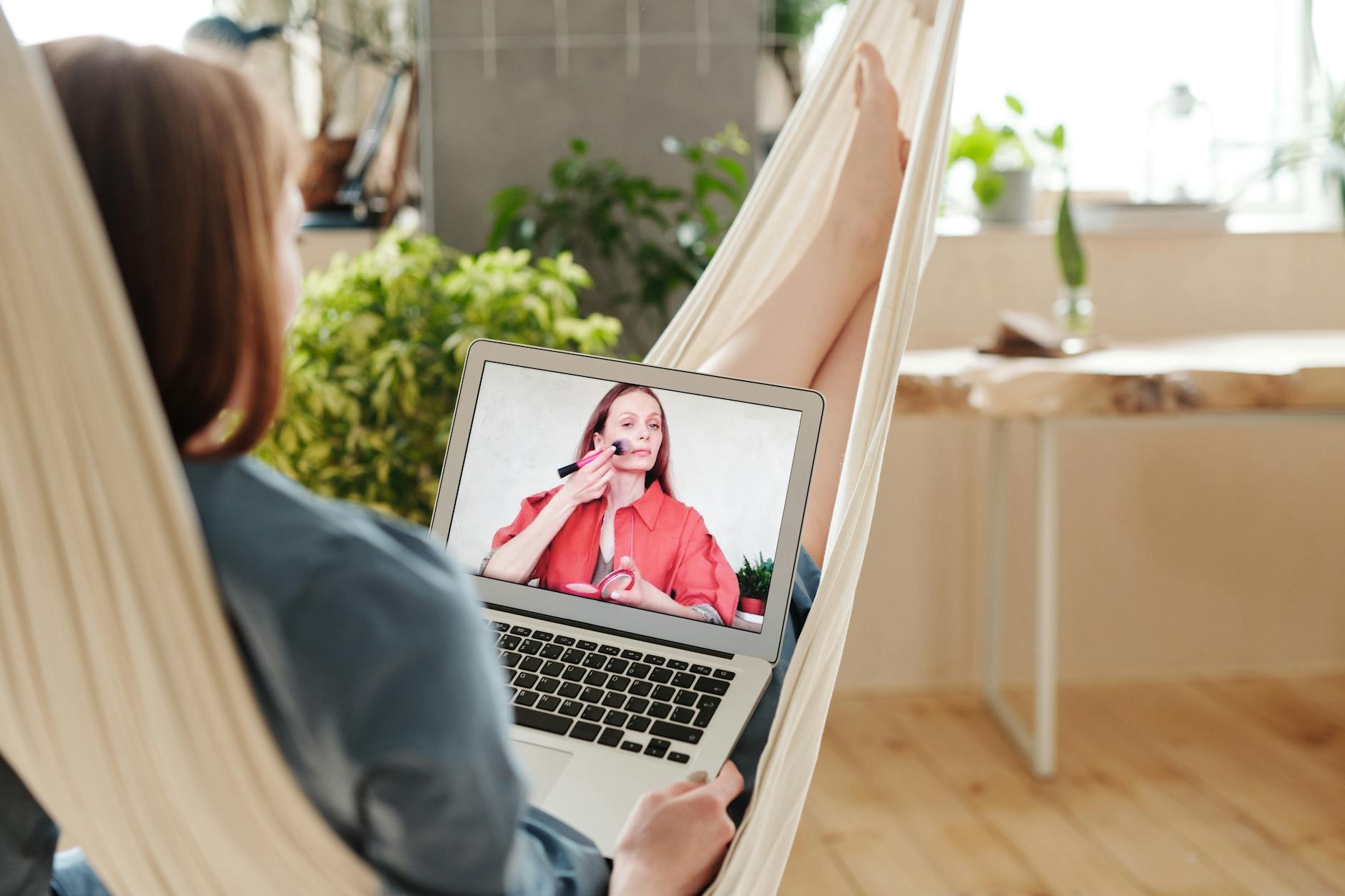 A woman comfortably lounges on a hammock indoors, watching a makeup tutorial on her laptop.