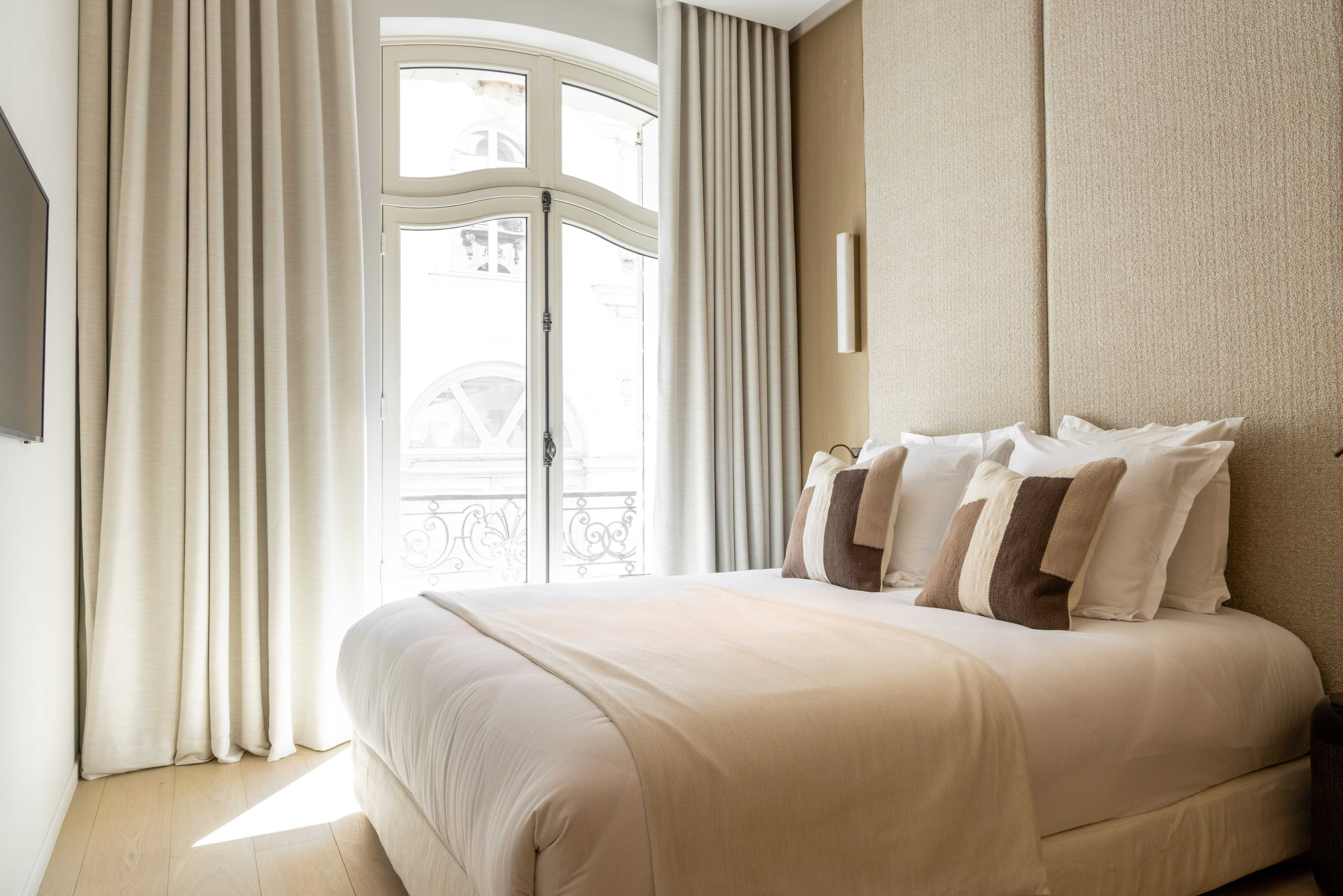 Chambre elegante avec lit double, coussins sable et bruns, tete de lit matelassee beige et fenetre cintre sur balcon parisien