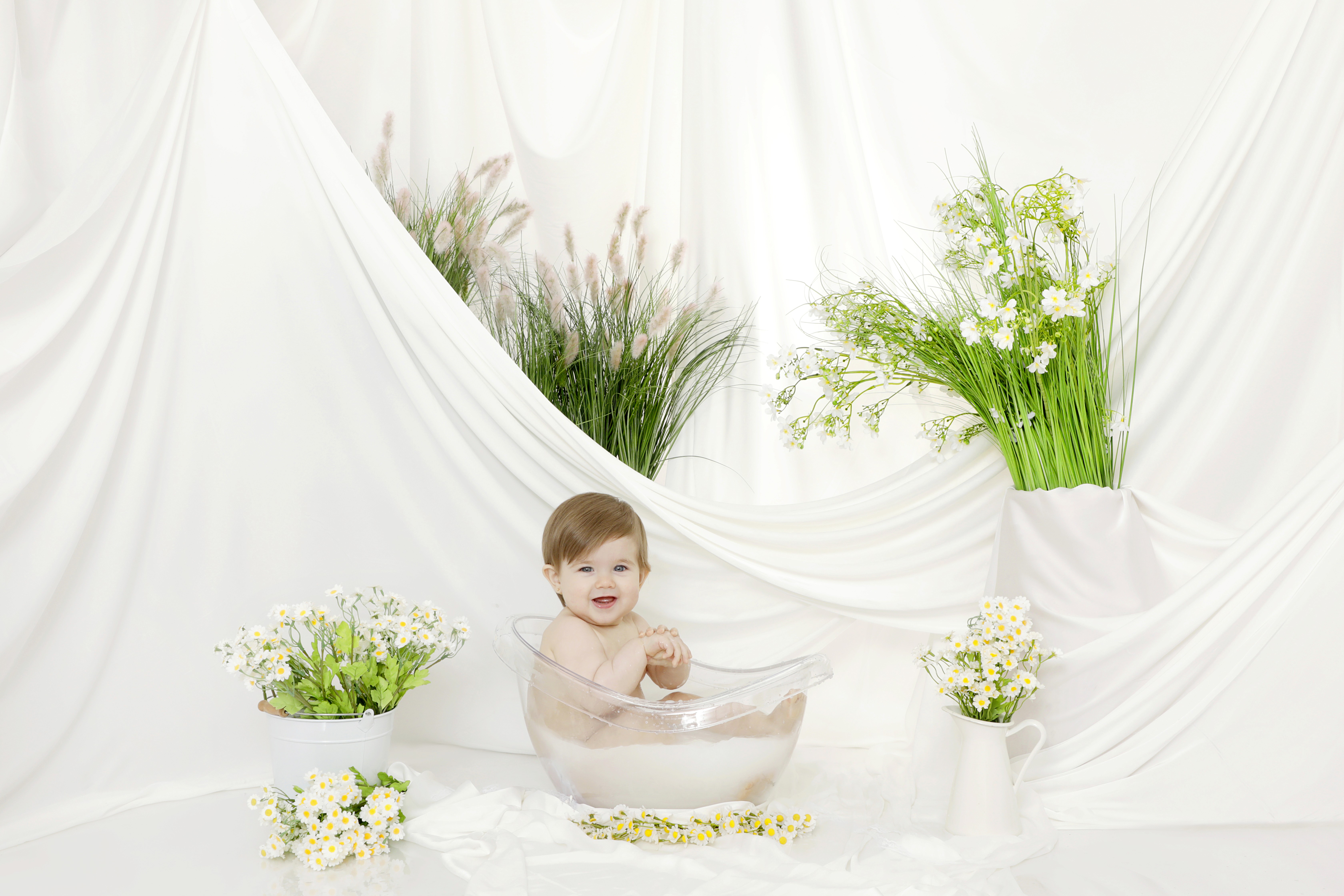A baby is sitting in a clear tub with milk, surrounded by white grapes, green plants, and small white flowers. 