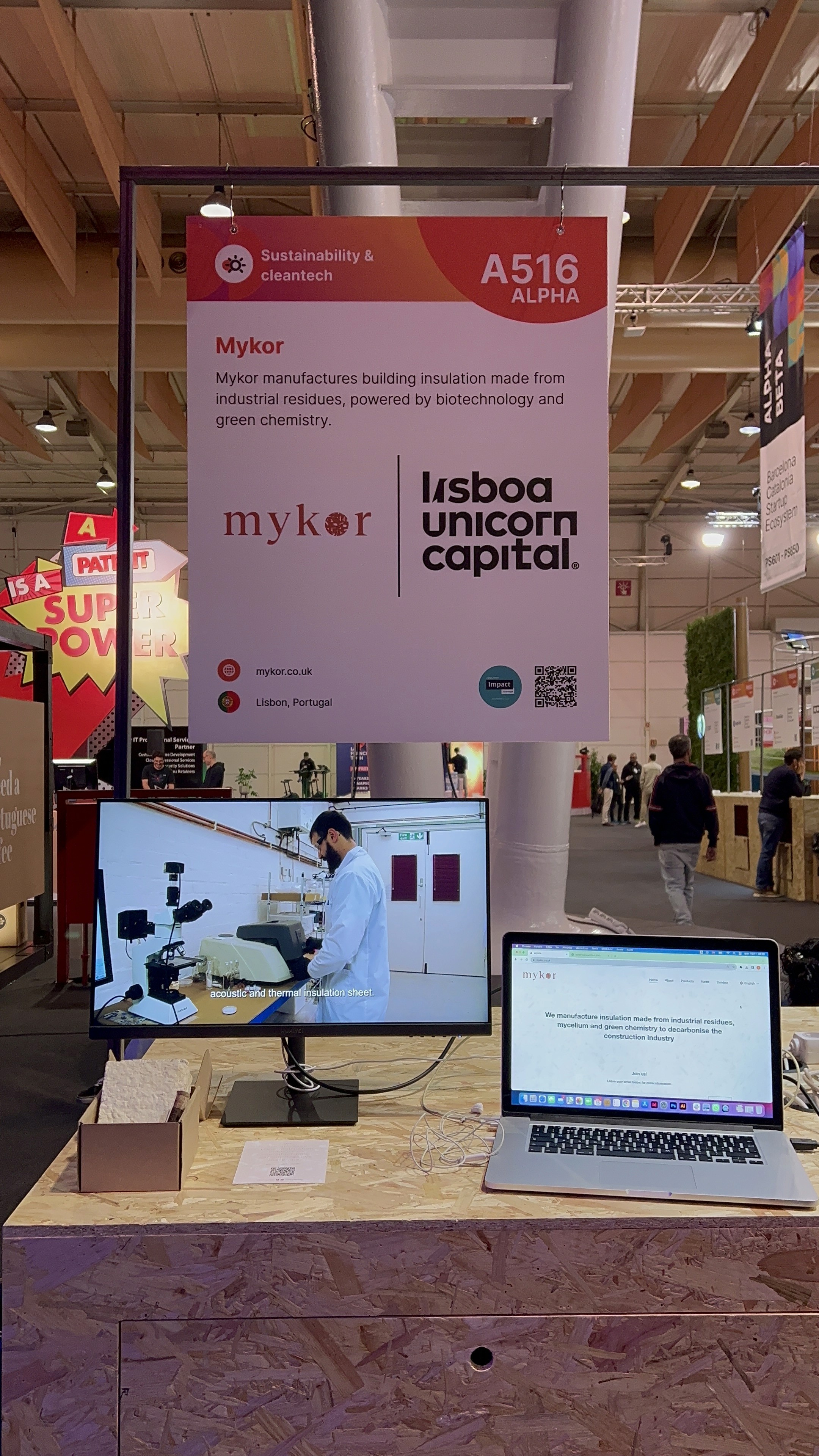 You can see an exhibition stand with a screen showing a video in a laboratory and a computer next to it showing Mykor's website. Behind those there is a banner with Mykor's information. 