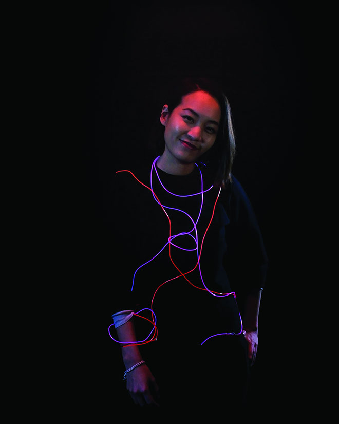 A young woman with dark hair stands against a black background. She wears a black long-sleeved shirt and has her arms relaxed at her sides. Colorful light trails—red, blue, and purple—form a swirling pattern across her torso and arms. She looks directly at the camera with a slight smile.