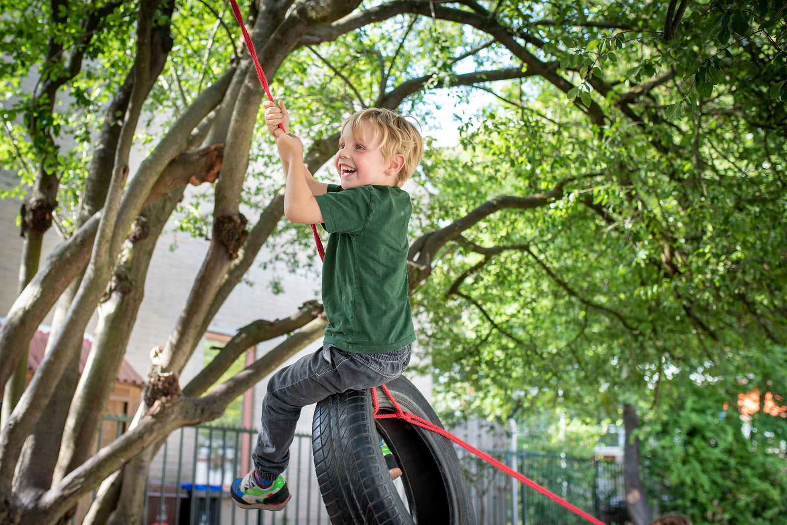 Een jonge jongen met blond haar en een groen T-shirt schommelt lachend op een autoband aan een rood touw in een groene, boomrijke speeltuin.