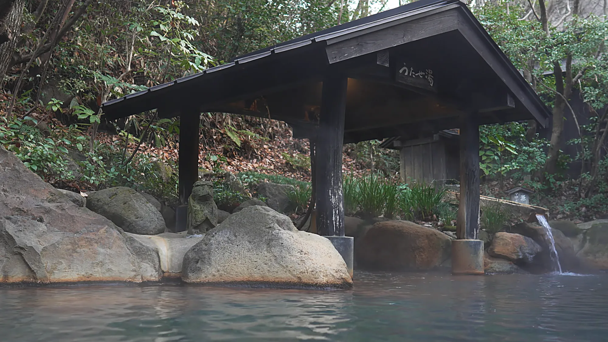 An open-air pool under a timber shelter, boulders and a small cascade behind