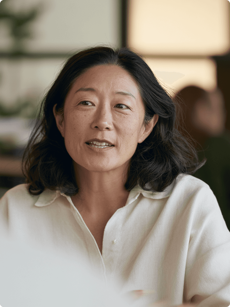 Woman in a white linen shirt in conversation across a table in a softly lit room."