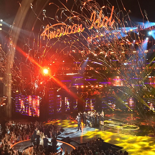 People standing on a brightly lit stage with "American Idol" sign above, surrounded by confetti and an applauding audience.