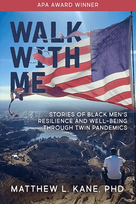 A young person with curly hair smiles at the camera while sitting in front of a laptop against a dark background.