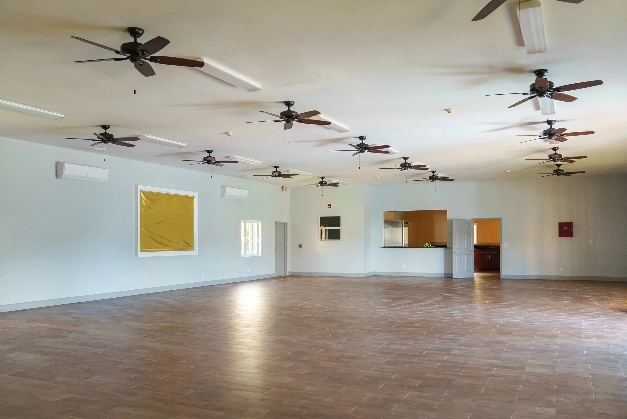 A large, empty room with a tiled floor and multiple ceiling fans. The walls are plain with a few small openings leading to other spaces. Bright lights illuminate the room, creating a spacious and airy atmosphere.