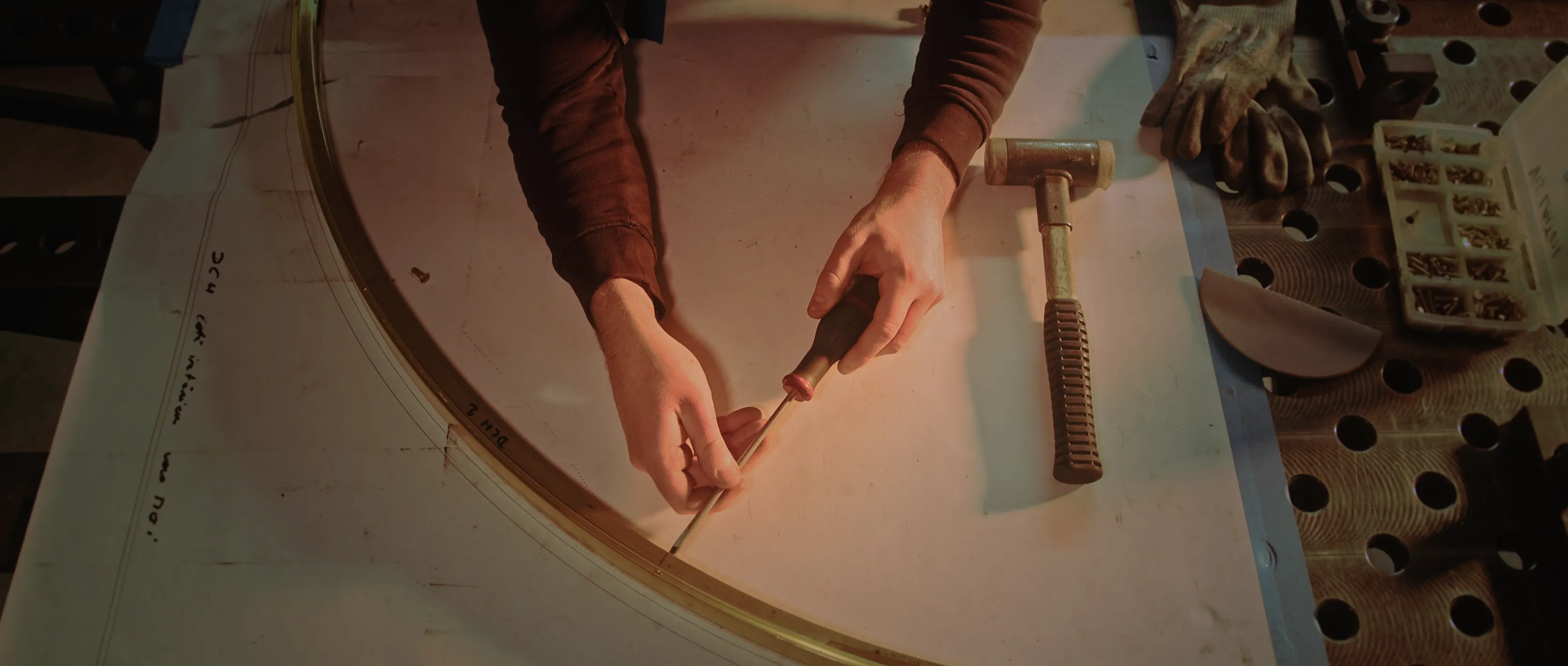 A person in a workshop meticulously crafts a curved metal piece using a hammer and a small tool, surrounded by precise measurements and tools on a perforated workbench.