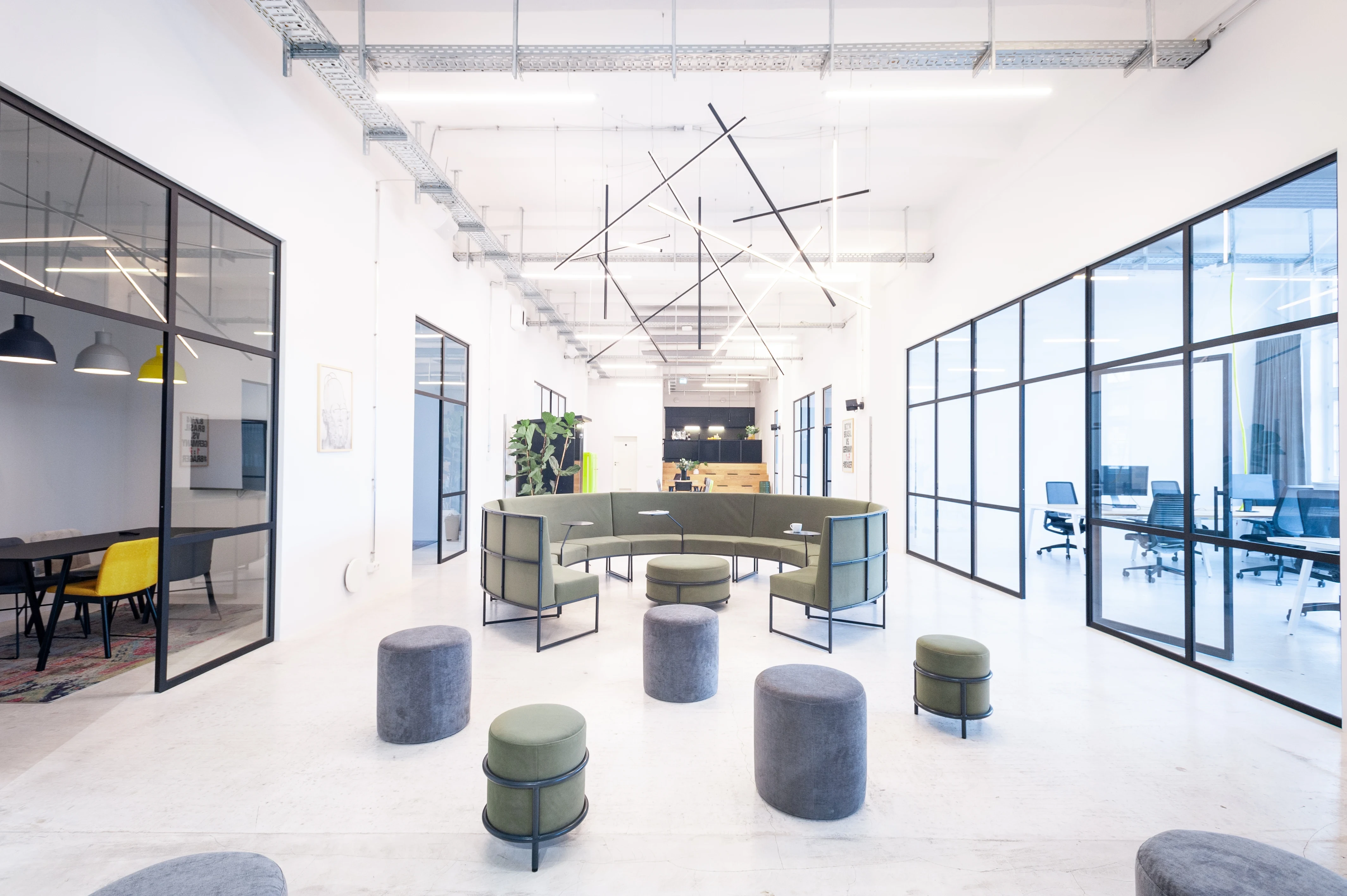 Open lounge area at LA RED with a curved green sofa, scattered grey and green poufs, black graphic pendant lights above and glass-walled offices on both sides.