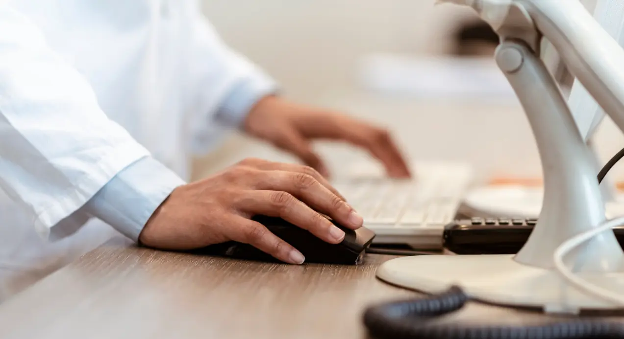 Mano di un operatore in camice bianco che usa mouse e tastiera al computer.