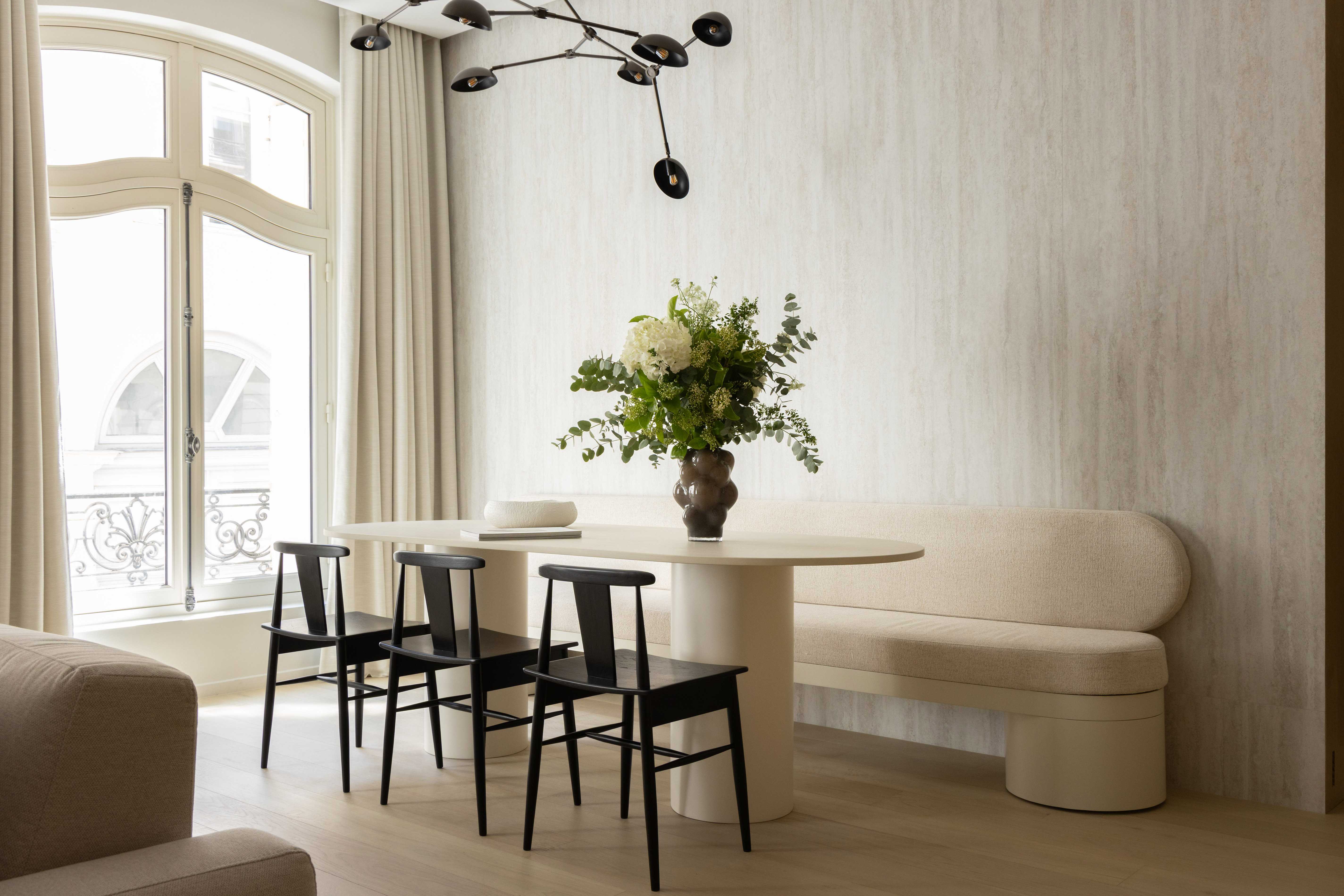 Salle a manger avec table ovale, banquette courbe beige, chaises noires et bouquet de fleurs pres de la fenetre cintre