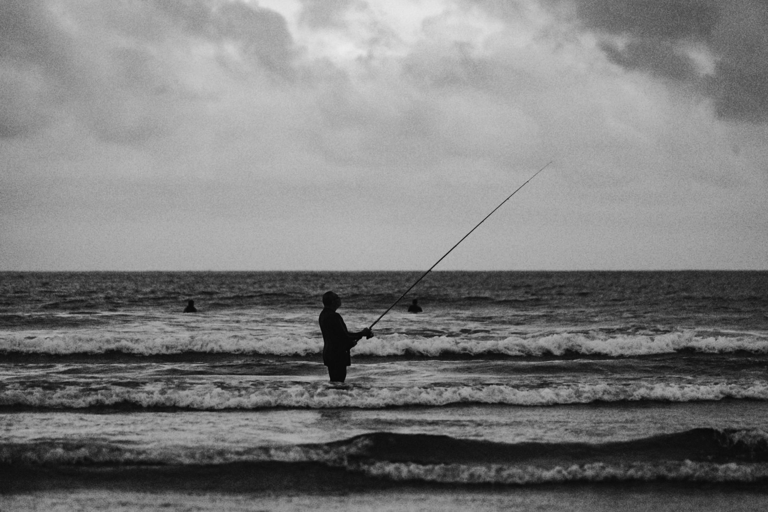 Black and white photo of a the beach on a cloudy day and a silhouette of someone fishing and two surfers in the water.