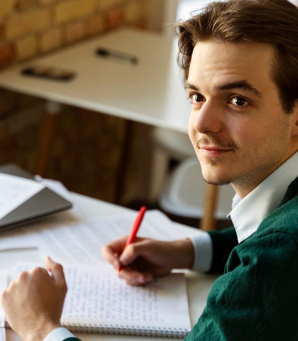 A person with short brown hair wearing a green sweater sits at a desk with scattered papers and a red pen in hand, against a backdrop of a brick wall and modern furniture. 