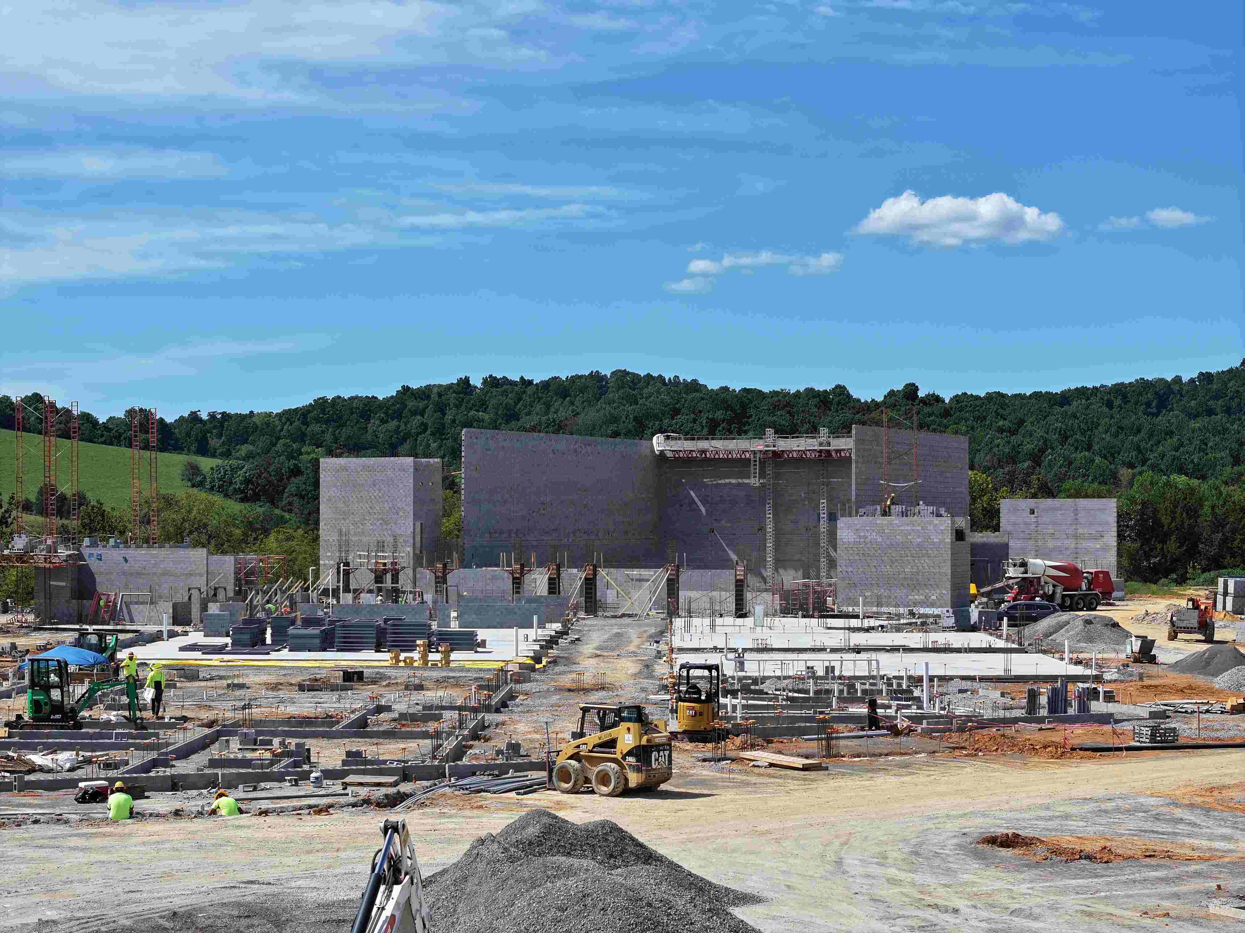 Construction site with machinery and materials in foreground, surrounded by lush green hills and a clear blue sky.