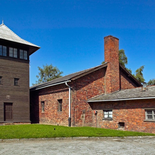 A brick building with a tall chimney stands next to a wooden tower under a clear blue sky. The scene includes grass and pavement.