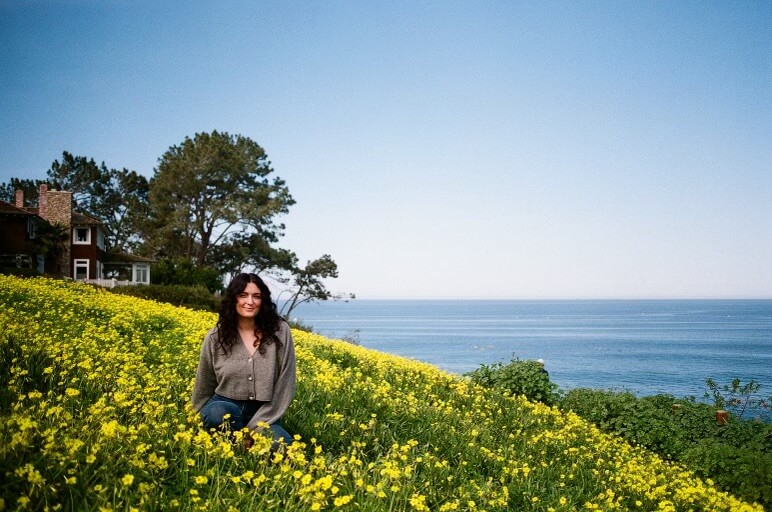 a film photo of Rae Cohen in a cliffside meadow of yellow flowers overlooking the ocean