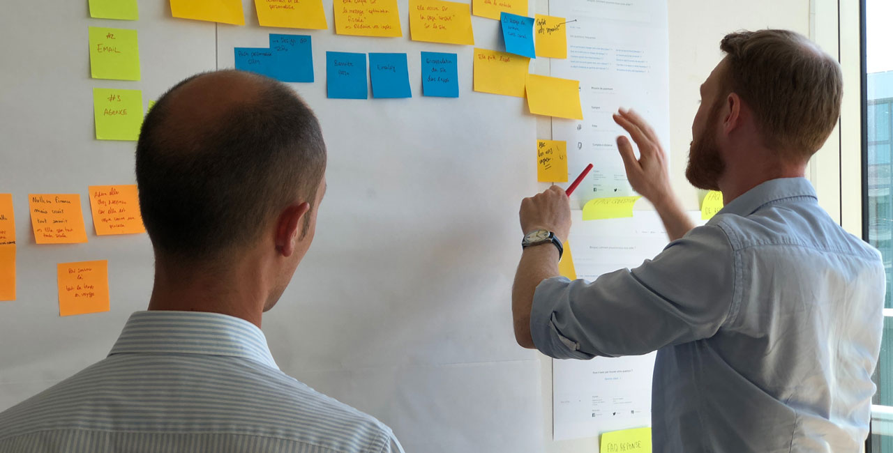 Two men organizing colorful sticky notes on a wall during a UX workshop brainstorming activity