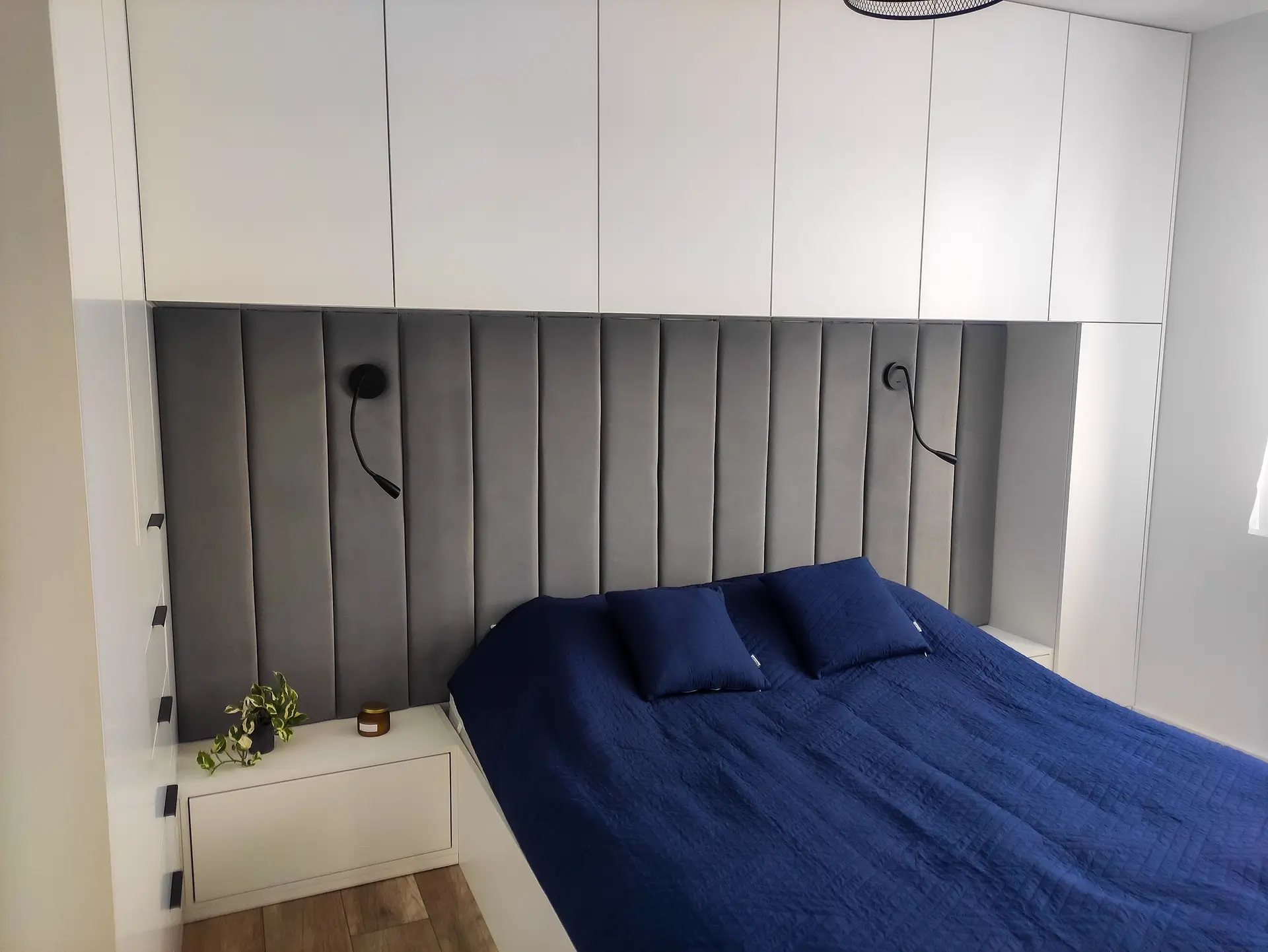 Nowoczesna sypialnia z tapicerowanym zagłówkiem w kolorze ciemnoszarym i białymi wiszącymi szafkami.