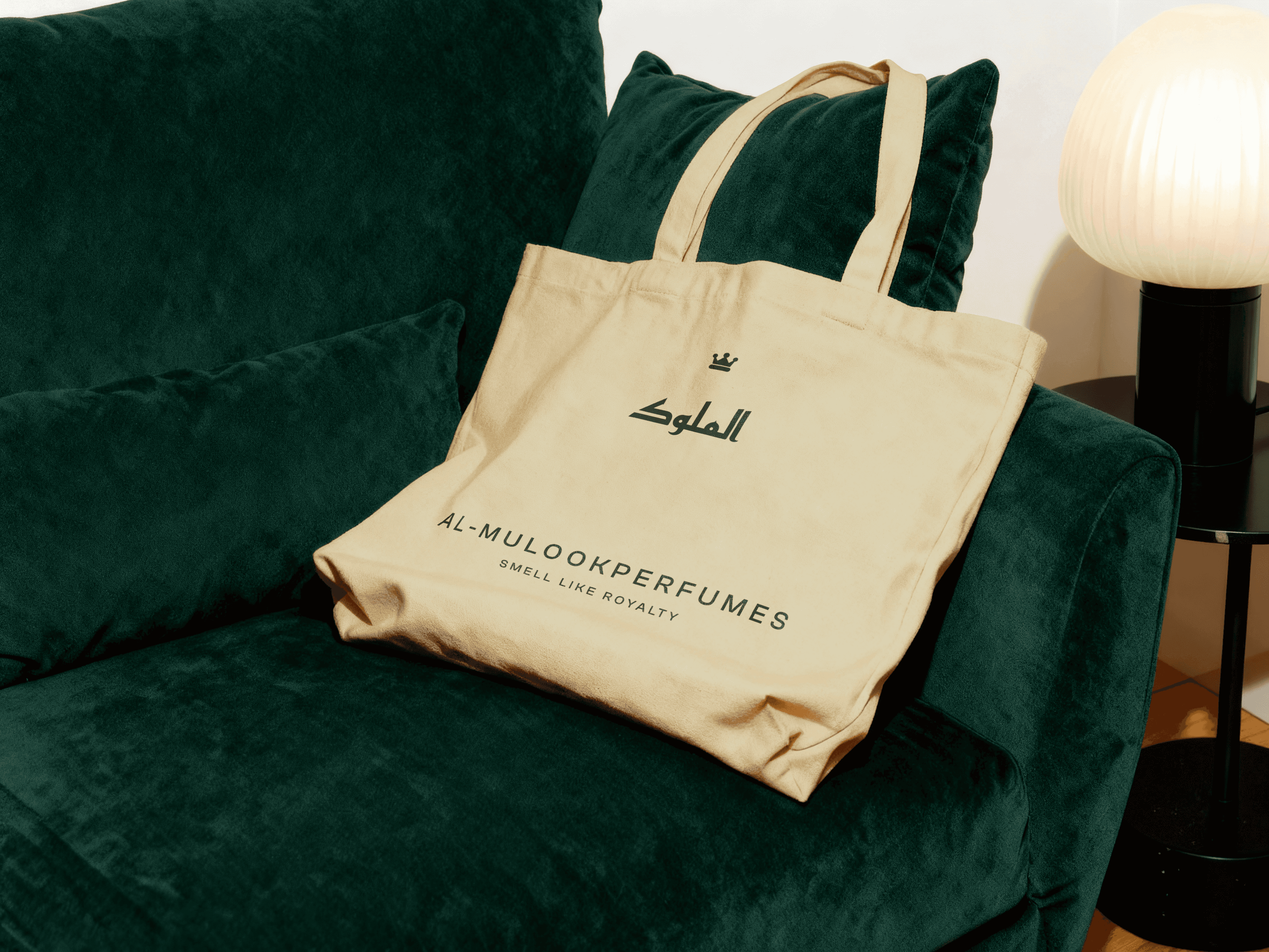 Beige Al-Mulook tote bag with Arabic logo and “Smell Like Royalty” tagline placed on a dark green velvet sofa beside a soft-glow table lamp.