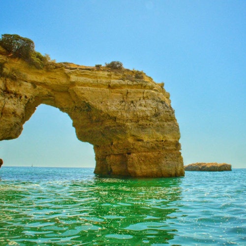Rock formation with a natural arch rising from turquoise seawater under a clear blue sky. A smaller rock is visible in the background.