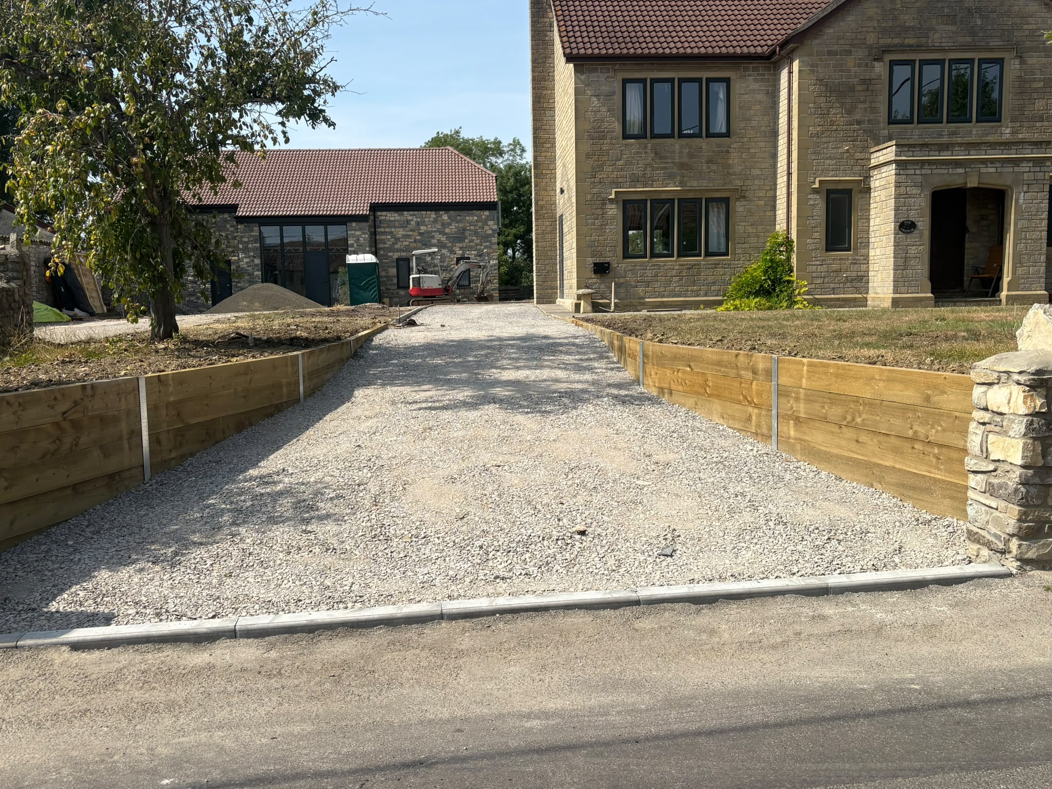 Gravel driveway leading to a stone house, bordered by timber retaining walls.