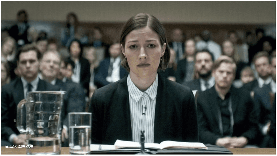 A woman sits solemnly at a hearing, facing a serious inquiry in a scene from the Black Mirror episode 'Hated in the Nation,' which explores themes of surveillance, social media, and justice.