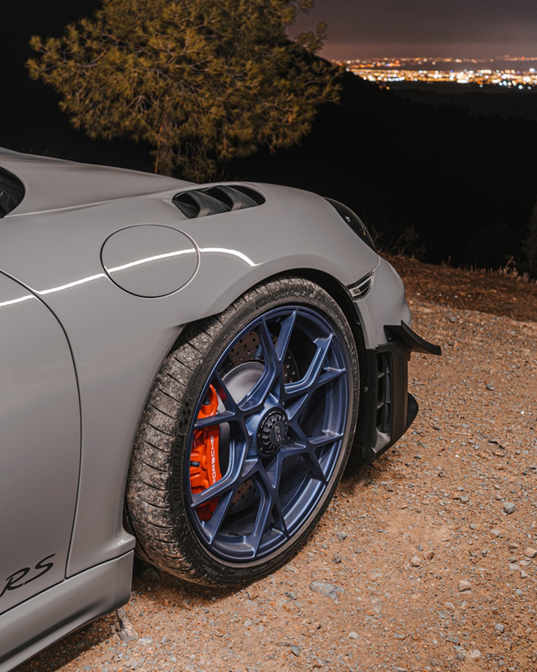 Close-up of a grey Porsche 718 Cayman GT4 RS front wheel, showing the blue multi-spoke rim, orange brake caliper and Michelin Pilot Sport Cup 2 tyre, with city lights glowing far below in the night.